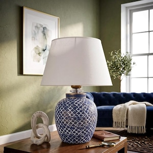 May include: A table lamp with a white tapered fabric shade and a rounded navy blue ceramic base featuring a white scalloped fish scale pattern. The base is accented with a clear glass detail at the neck and sits on a dark wooden surface.