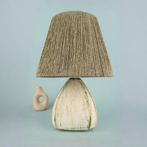 May include: A table lamp featuring a beige rope shade and a white, teardrop-shaped base with green detailing. A small, off-white vase with a circular design is positioned beside the lamp. The background is a light blue colour.