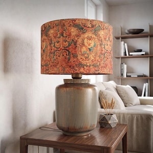 May include: A table lamp with a cylindrical, weathered grey base and a drum-shaped lampshade featuring a detailed, multi-coloured floral and scrollwork pattern in shades of orange, red, green, and gold, set on a dark wooden side table.