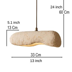 K&ouml;nnte beinhalten: Beige, l&auml;ngliche Pendelleuchte. Die Leuchte ist 33 cm lang, 13 cm breit und h&auml;ngt 60 cm von der Decke. Die Leuchte hat eine strukturierte Oberfl&auml;che und ein messingfarbenes Baldachin.