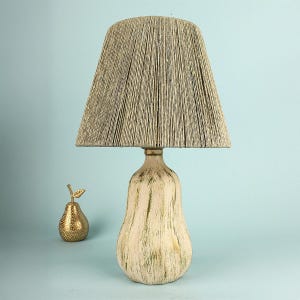 May include: A table lamp featuring a beige, textured lampshade and a pear-shaped base. The base is off-white with green accents. A gold-coloured pear figurine sits beside the lamp, adding a touch of elegance to the decor.