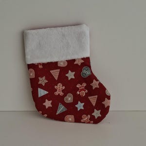 May include: A Christmas stocking featuring a deep red fabric base with a repeating pattern of gingerbread people, stars, hearts, and triangular trees in white and light blue. The top cuff is made of a thick, white, plush faux fur material.