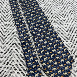 May include: A navy blue tie featuring a repeating pattern of cream elephants and orange floral motifs. The tie is made of a smooth material and is displayed against a textured black and white background. A classic accessory for formal or business wear.