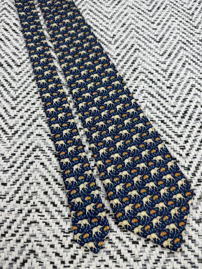 May include: A navy blue tie featuring a repeating pattern of cream elephants and orange floral motifs. The tie is made of a smooth material and is displayed against a textured black and white background. A classic accessory for formal or business wear.