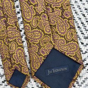 May include: A necktie featuring a brown background with a repeating yellow and purple paisley pattern. The back of the tie shows a dark blue lining with a rectangular label that reads Jim Thompson in white text, indicating the brand of the accessory.