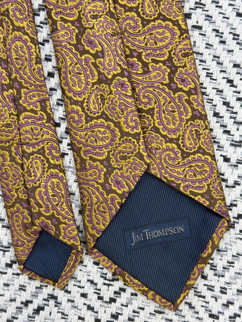 May include: A necktie featuring a brown background with a repeating yellow and purple paisley pattern. The back of the tie shows a dark blue lining with a rectangular label that reads Jim Thompson in white text, indicating the brand of the accessory.