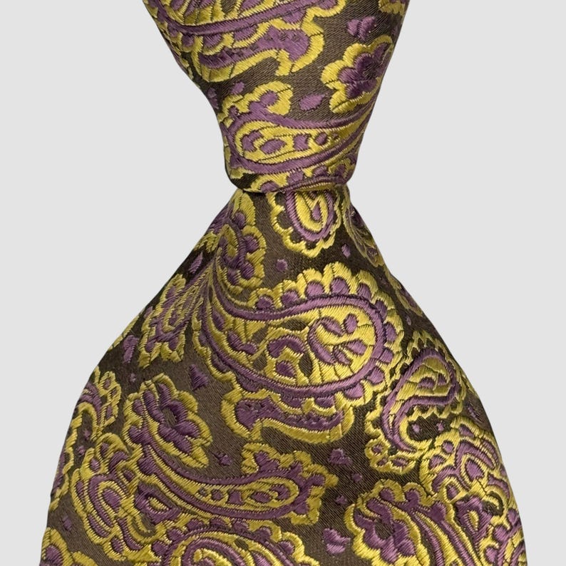 May include: A necktie featuring a dark brown base color with an intricate, repeating paisley pattern in shades of vibrant yellow and muted purple. The fabric has a subtle sheen, and the tie is knotted at the top, displaying the detailed woven design.