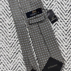 May include: A Jim Thompson tie with a black, white, and tan checkered pattern. The tie features the Jim Thompson brand name on the label and the back of the tie. The tie is displayed on a textured, black and white background.
