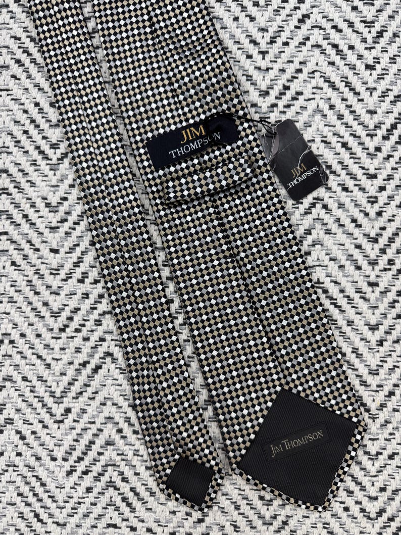 May include: A Jim Thompson tie with a black, white, and tan checkered pattern. The tie features the Jim Thompson brand name on the label and the back of the tie. The tie is displayed on a textured, black and white background.