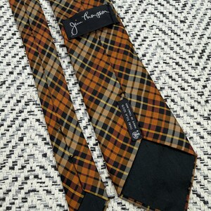 Puede incluir: Una corbata Jim Thompson con un patr&oacute;n de cuadros en tonos naranja, negro y blanco. La parte posterior de la corbata incluye una etiqueta negra con el nombre de la marca y una segunda etiqueta negra que indica Thai Silk 100% Made in Thailand.