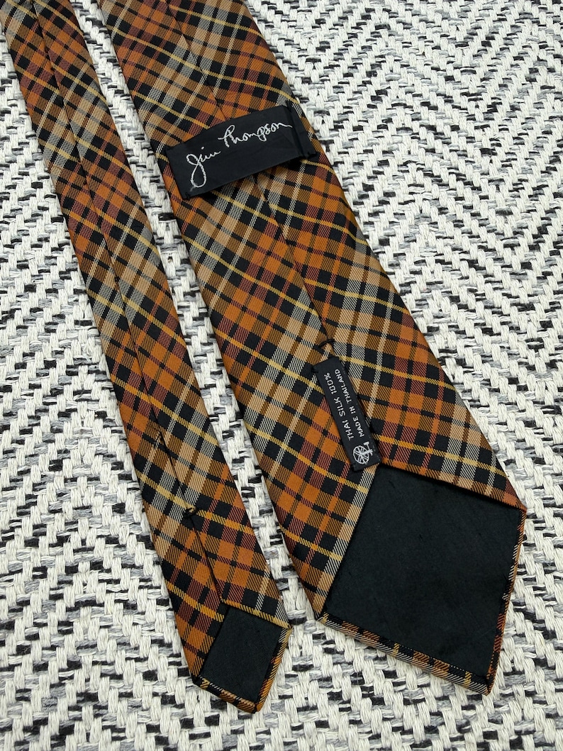 Puede incluir: Una corbata Jim Thompson con un patr&oacute;n de cuadros en tonos naranja, negro y blanco. La parte posterior de la corbata incluye una etiqueta negra con el nombre de la marca y una segunda etiqueta negra que indica Thai Silk 100% Made in Thailand.