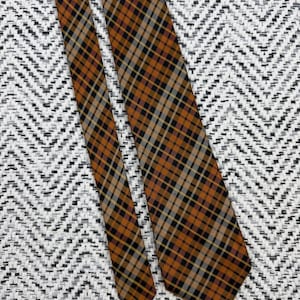 Puede incluir: Una corbata con un patr&oacute;n de cuadros en diagonal en tonos naranja quemado, negro, beige y gris. La tela tiene una textura tejida con una punta afilada y un extremo m&aacute;s estrecho, mostrando una cuadr&iacute;cula consistente de l&iacute;neas cruzadas.