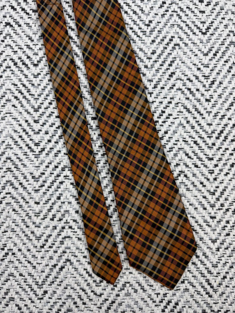 Puede incluir: Una corbata con un patr&oacute;n de cuadros en diagonal en tonos naranja quemado, negro, beige y gris. La tela tiene una textura tejida con una punta afilada y un extremo m&aacute;s estrecho, mostrando una cuadr&iacute;cula consistente de l&iacute;neas cruzadas.