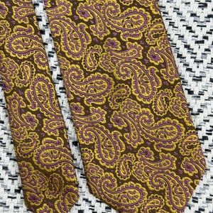 May include: A necktie featuring a dark brown background with a repeating paisley pattern in mustard yellow and muted purple. The fabric has a visible woven texture, and the design covers the entire surface of the tie in a consistent, dense arrangement.