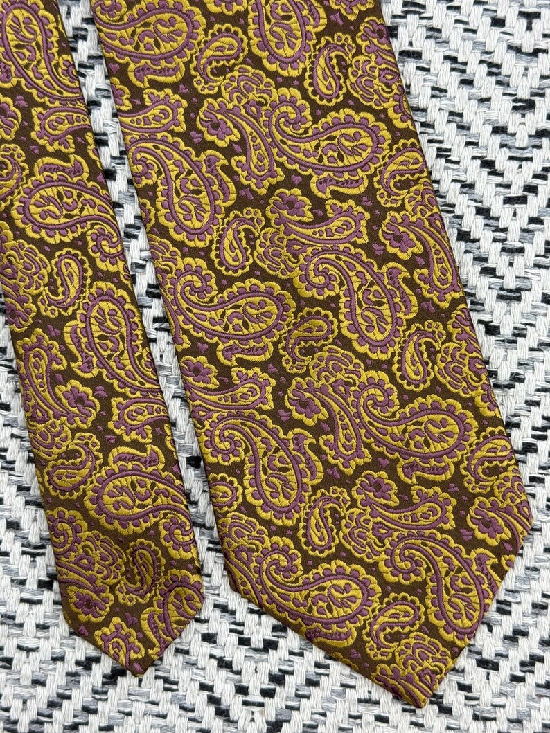 May include: A necktie featuring a dark brown background with a repeating paisley pattern in mustard yellow and muted purple. The fabric has a visible woven texture, and the design covers the entire surface of the tie in a consistent, dense arrangement.