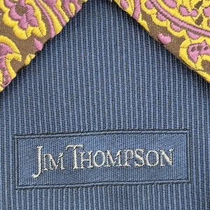 May include: A necktie featuring a brown fabric with a yellow and purple paisley pattern. The reverse side displays a blue and black pinstriped lining with a rectangular label stitched in the center that reads Jim Thompson in white lettering.
