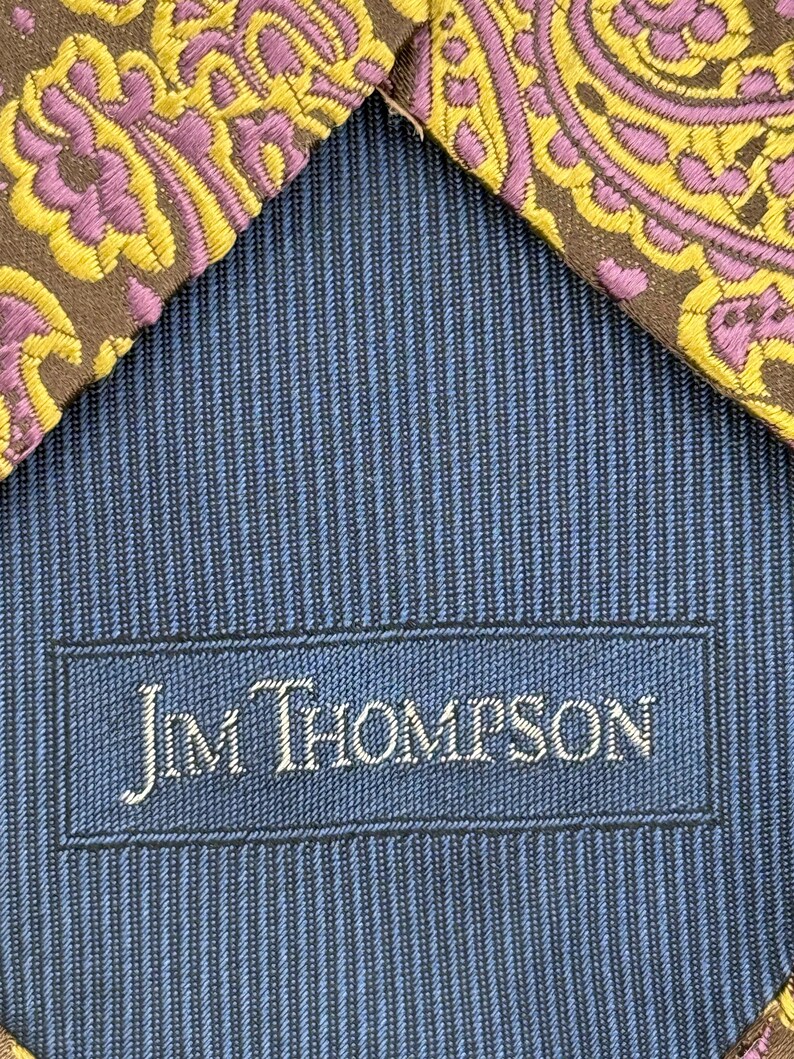 May include: A necktie featuring a brown fabric with a yellow and purple paisley pattern. The reverse side displays a blue and black pinstriped lining with a rectangular label stitched in the center that reads Jim Thompson in white lettering.