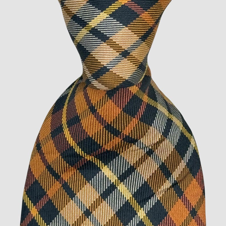 Puede incluir: Una corbata con un patr&oacute;n de cuadros tejido en tonos azul marino oscuro, amarillo mostaza, naranja quemado y blanco. La tela tiene una textura diagonal visible y el dise&ntilde;o consiste en l&iacute;neas que se cruzan formando una cuadr&iacute;cula cl&aacute;sica.