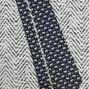 May include: A navy blue tie featuring a repeating pattern of cream elephants and orange floral details. The tie is displayed on a black and white chevron patterned surface.