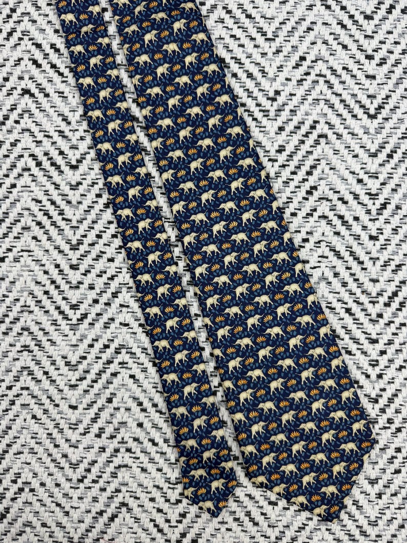 May include: A navy blue tie featuring a repeating pattern of cream elephants and orange floral details. The tie is displayed on a black and white chevron patterned surface.