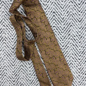 May include: A necktie featuring a brown base color with an intricate paisley pattern in shades of mustard yellow and purple. The tie is folded, showing both the wide blade and the narrower tail end with a pointed tip and a smooth fabric texture.