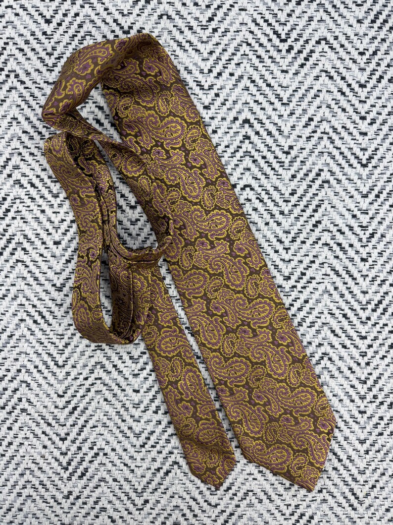 May include: A necktie featuring a brown base color with an intricate paisley pattern in shades of mustard yellow and purple. The tie is folded, showing both the wide blade and the narrower tail end with a pointed tip and a smooth fabric texture.
