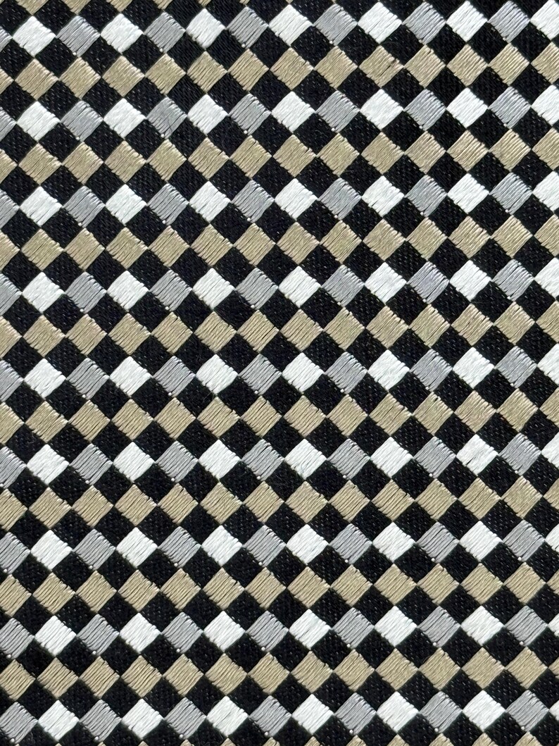 May include: Close-up of a fabric with a repeating diamond pattern. The diamonds are in black, white, silver, and tan. The fabric has a textured appearance, suggesting a woven or embroidered design. The pattern is symmetrical and creates a visually interesting surface.