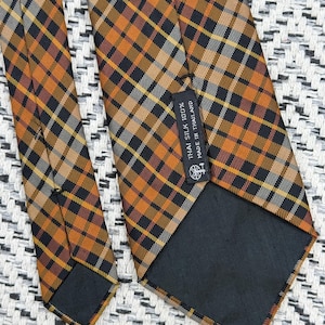 Puede incluir: Una corbata con un patr&oacute;n de cuadros en tonos naranja, negro y blanco. La parte posterior de la corbata incluye un forro de tela negra y una etiqueta rectangular con el texto: THAI SILK 100% MADE IN THAILAND, junto a un peque&ntilde;o logotipo.