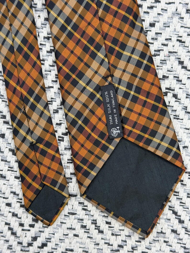 Puede incluir: Una corbata con un patr&oacute;n de cuadros en tonos naranja, negro y blanco. La parte posterior de la corbata incluye un forro de tela negra y una etiqueta rectangular con el texto: THAI SILK 100% MADE IN THAILAND, junto a un peque&ntilde;o logotipo.
