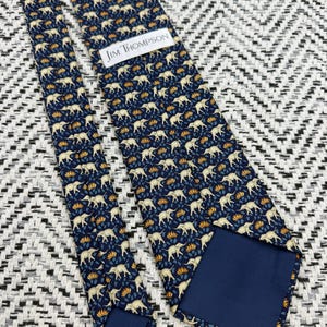 May include: A navy blue tie featuring a repeating pattern of cream elephants and orange and blue floral details. The label reads "JIM THOMPSON". The tie is displayed on a black and white patterned surface.