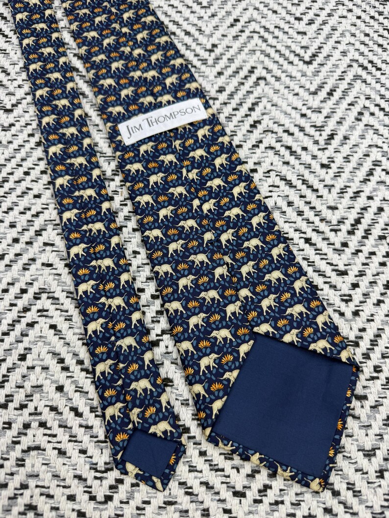 May include: A navy blue tie featuring a repeating pattern of cream elephants and orange and blue floral details. The label reads "JIM THOMPSON". The tie is displayed on a black and white patterned surface.