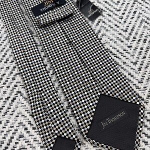 May include: A Jim Thompson brand necktie with a checkered pattern in black, white, and tan. The tie is made of a smooth fabric and has a black backing. The Jim Thompson label is visible on the front and back of the tie.