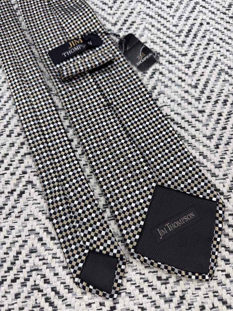 May include: A Jim Thompson brand necktie with a checkered pattern in black, white, and tan. The tie is made of a smooth fabric and has a black backing. The Jim Thompson label is visible on the front and back of the tie.
