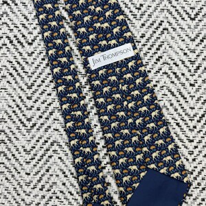May include: A navy blue silk tie with a repeating pattern of cream elephants and yellow floral accents. The tie features the text "JIM THOMPSON" on a white label.