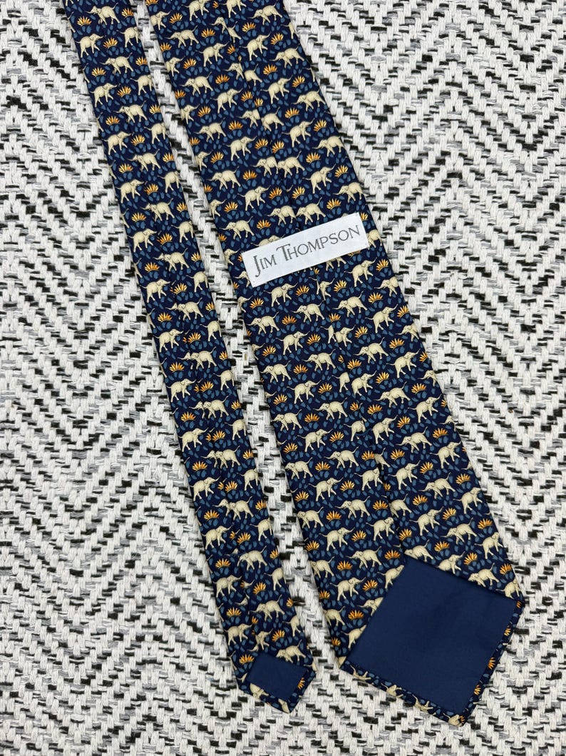 May include: A navy blue silk tie with a repeating pattern of cream elephants and yellow floral accents. The tie features the text "JIM THOMPSON" on a white label.