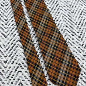 Puede incluir: Una corbata con un patr&oacute;n de cuadros en tonos naranja quemado, negro, blanco y gris. La tela muestra un dise&ntilde;o de cuadr&iacute;cula diagonal con l&iacute;neas que se cruzan de diferentes grosores, creando una apariencia geom&eacute;trica estructurada.