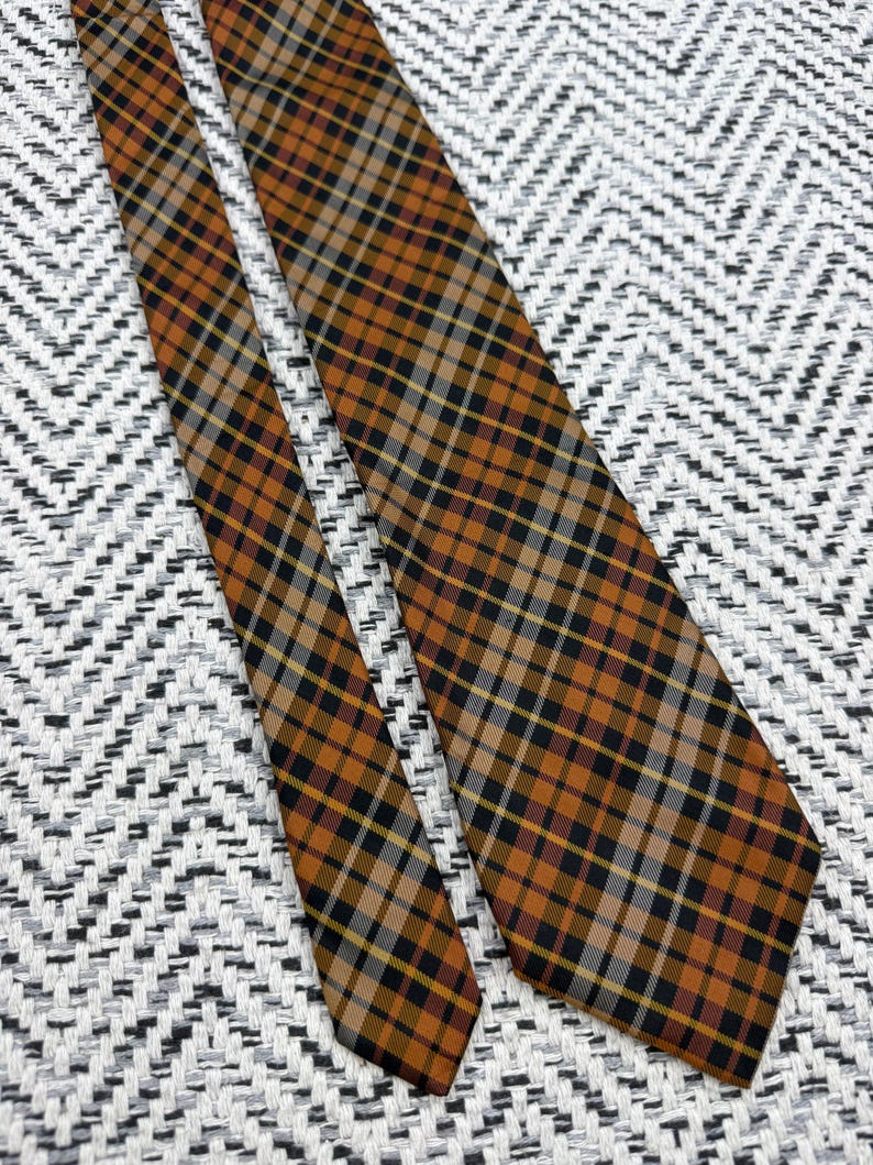 Puede incluir: Una corbata con un patr&oacute;n de cuadros en tonos naranja quemado, negro, blanco y gris. La tela muestra un dise&ntilde;o de cuadr&iacute;cula diagonal con l&iacute;neas que se cruzan de diferentes grosores, creando una apariencia geom&eacute;trica estructurada.