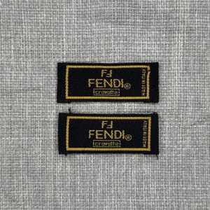 May include: Two rectangular black fabric labels with gold embroidered borders and text. The text reads Fendi, with the Fendi logo above, the word cravatte below, and Made in Italy printed vertically along the right side of each label.