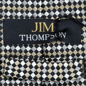 May include: Close-up of a patterned necktie with a black, silver, and tan checkered design. The label reads "JIM THOMPSON" in gold and white lettering. The tie is made of a woven fabric.
