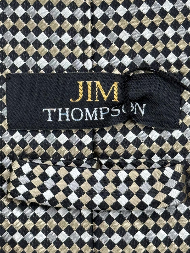 May include: Close-up of a patterned necktie with a black, silver, and tan checkered design. The label reads "JIM THOMPSON" in gold and white lettering. The tie is made of a woven fabric.