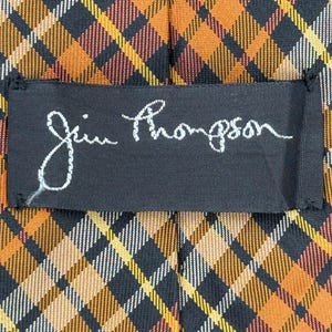 Puede incluir: Una corbata con un patr&oacute;n de cuadros en diagonal en tonos naranja, negro, amarillo y blanco. Una etiqueta rectangular de tela negra est&aacute; cosida en el centro, mostrando el texto bordado en blanco de la firma Jim Thompson.
