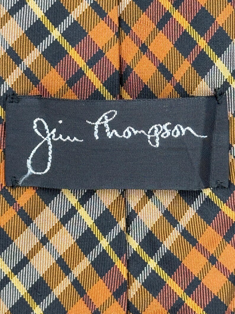 Puede incluir: Una corbata con un patr&oacute;n de cuadros en diagonal en tonos naranja, negro, amarillo y blanco. Una etiqueta rectangular de tela negra est&aacute; cosida en el centro, mostrando el texto bordado en blanco de la firma Jim Thompson.