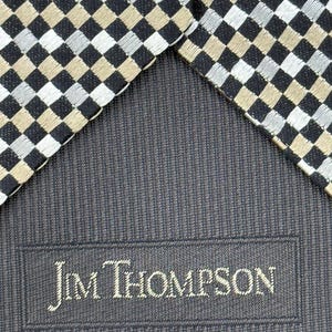 May include: Close-up of a necktie with a checkered pattern in black, silver, and tan. The tie's reverse side is a dark blue fabric with the embroidered text "Jim Thompson".