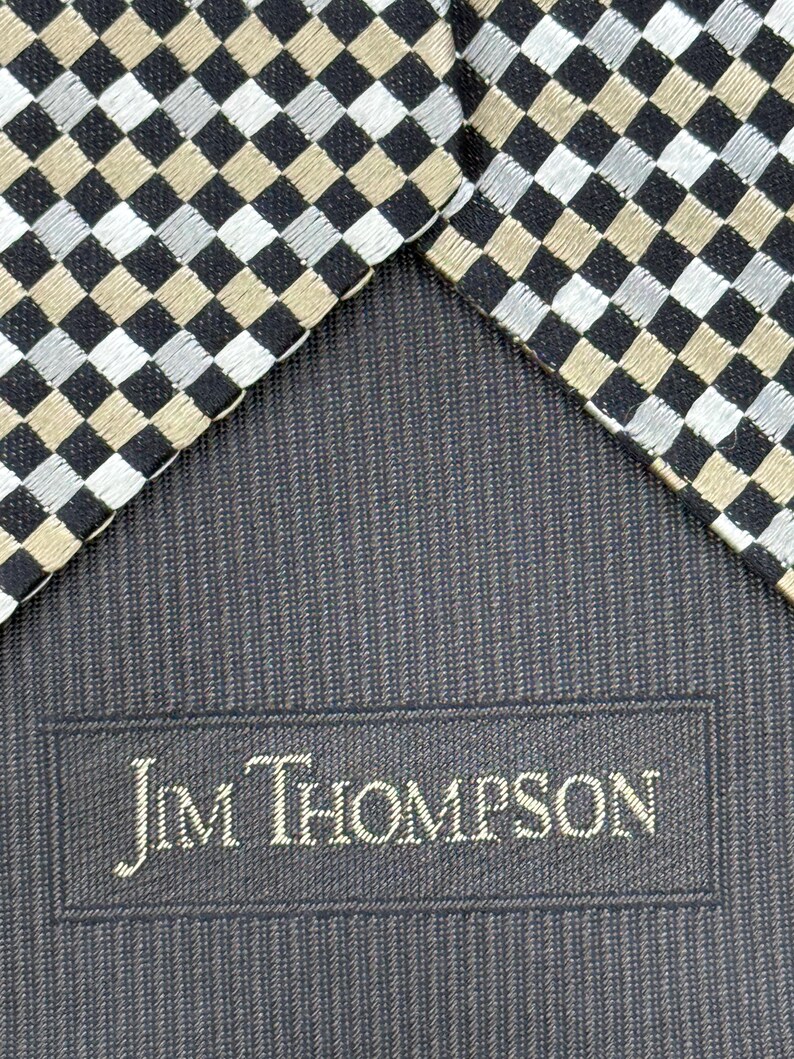 May include: Close-up of a necktie with a checkered pattern in black, silver, and tan. The tie's reverse side is a dark blue fabric with the embroidered text "Jim Thompson".