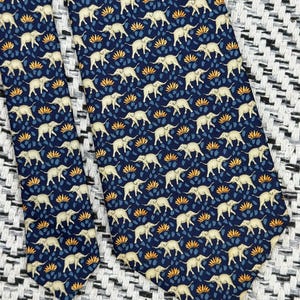 May include: Navy blue tie with a repeating pattern of cream elephants and orange and blue floral motifs. The tie is made of a smooth fabric, suitable for formal or business wear.