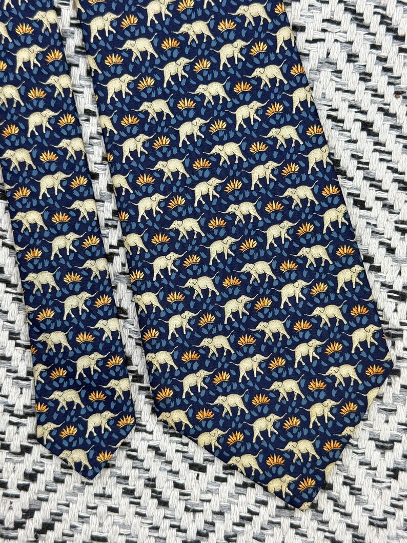 May include: Navy blue tie with a repeating pattern of cream elephants and orange and blue floral motifs. The tie is made of a smooth fabric, suitable for formal or business wear.