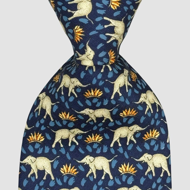 May include: A navy blue tie featuring a repeating pattern of light beige elephants, gold and blue floral motifs. The tie is knotted, highlighting the intricate design. A smart accessory for formal or business wear. The tie's length is approximately 147 cm.