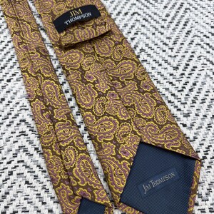 May include: A brown necktie featuring a repeating yellow and purple paisley pattern. The tie includes a black label with the text JIM THOMPSON stitched near the top and a dark blue fabric lining at the bottom with the text JIM THOMPSON printed on it.