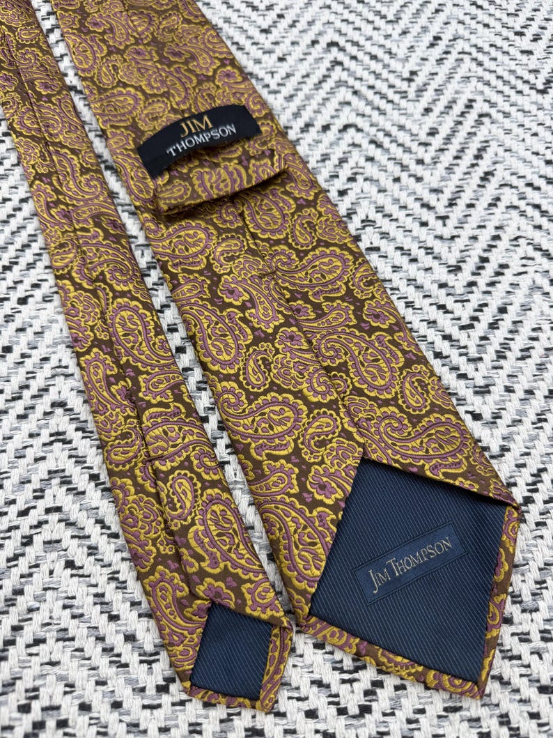 May include: A brown necktie featuring a repeating yellow and purple paisley pattern. The tie includes a black label with the text JIM THOMPSON stitched near the top and a dark blue fabric lining at the bottom with the text JIM THOMPSON printed on it.