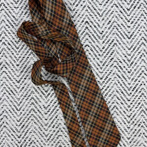 Puede incluir: Una corbata con un patr&oacute;n de cuadros en tonos naranja quemado, negro y beige. La tela tiene una textura suave y el dise&ntilde;o consiste en l&iacute;neas diagonales que se cruzan formando un motivo de cuadr&iacute;cula cl&aacute;sico a lo largo de todo el accesorio.